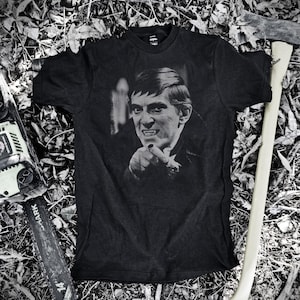 May include: Black t-shirt featuring a black and white image of a man pointing with a menacing expression. The image is surrounded by a chainsaw and an axe. The shirt is a dark color and appears to be made of cotton.