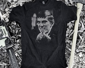 May include: Dark grey t-shirt with a black and white image of a man pointing. The image is surrounded by a chainsaw and an axe. The shirt is a dark colour and appears to be made of cotton. The image has a vintage feel.