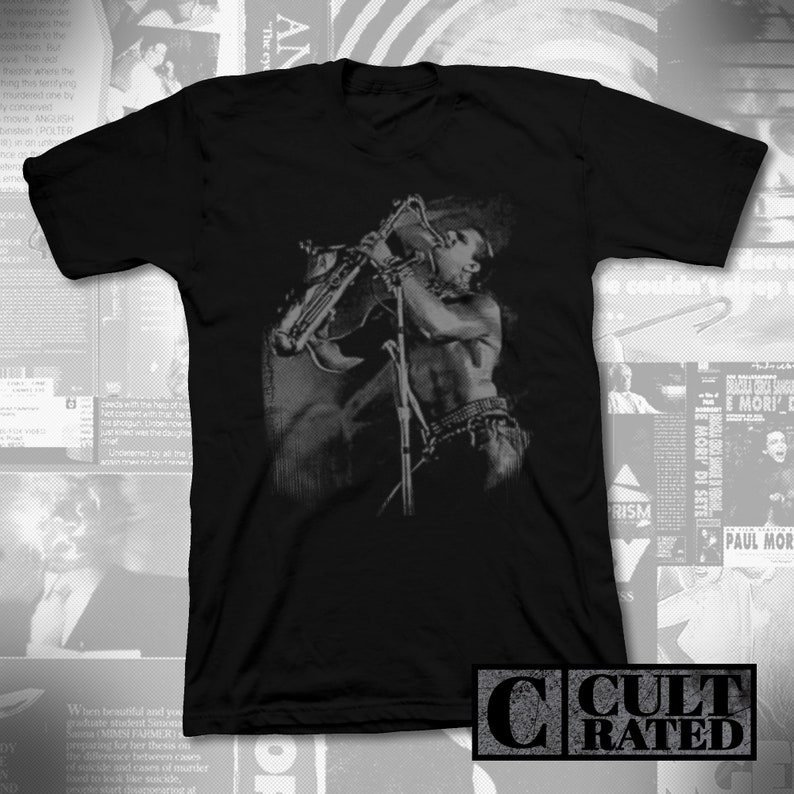May include: Black t-shirt with a faded gray image of a person playing a saxophone. The image is in a vintage style and has a grunge aesthetic. The t-shirt has a crew neck and short sleeves. The text "CULT RATED" is printed on the bottom right corner of the t-shirt.