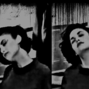 May include: Two women with dark hair and dark clothing pose for a black and white portrait. The women are looking down and their faces are partially obscured by shadows.