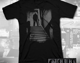 May include: Black t-shirt featuring a grey graphic print of a figure holding a chainsaw and standing on a staircase. The text "CULT RATED" is printed in white on a black background at the bottom of the image.