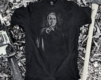 May include: Dark grey t-shirt with a grayscale image of a man pointing. A chainsaw and an axe are positioned near the shirt. The shirt rests on a bed of leaves and twigs, creating a rustic, horror-themed aesthetic.