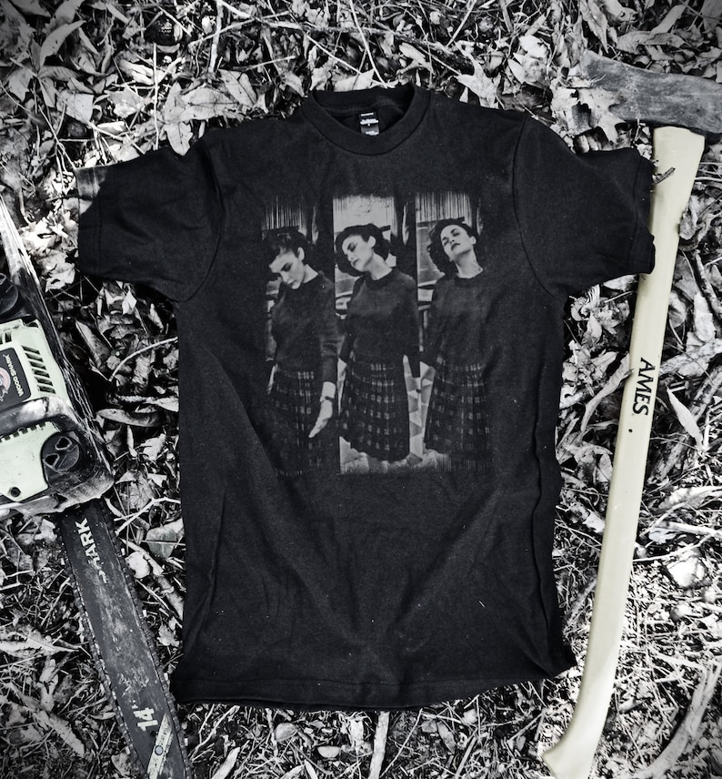 May include: Black t-shirt with a graphic print of three women wearing plaid skirts. The graphic is in black and white and the text "In the Pines" is printed below the image.