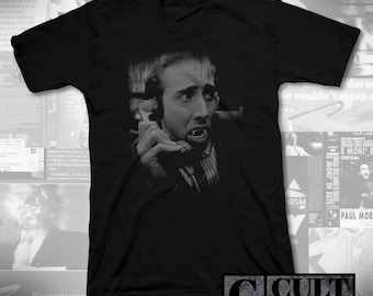 May include: Black V-neck t-shirt with a black and white image of a man with vampire fangs on the phone. The text "CULT RATED" is in the lower right corner. The image is a movie still.