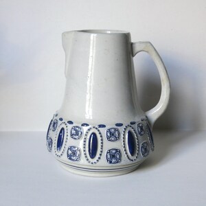 Può includere: Brocca in ceramica bianca con un motivo geometrico blu e bianco. La brocca ha una base larga e un corpo arrotondato. Il manico è attaccato al lato della brocca.