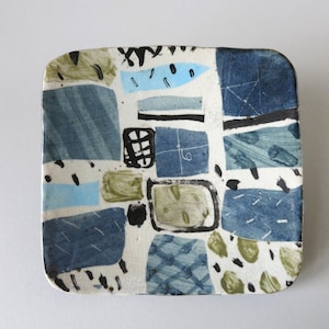 Op de afbeelding: Een vierkante keramische schaal met een witte basis, versierd met diverse abstracte vormen in tinten blauw, olijfgroen en zwart. Het oppervlak toont een mix van effen blokken, kruisarceringen en verspreide penseelstreekpatronen.