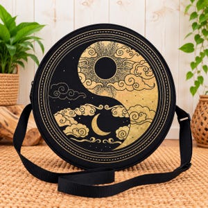 May include: A round black crossbody bag featuring a gold-toned yin and yang design. The graphic depicts a sun and clouds on one side and a crescent moon with clouds on the other, all enclosed within a circular border with a black strap.
