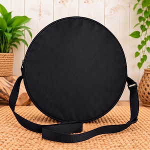 May include: A solid black circular crossbody bag featuring a smooth, matte fabric exterior. The bag includes an adjustable black nylon shoulder strap attached to the sides with silver-toned metal hardware, creating a simple, functional design.