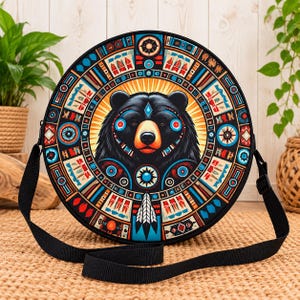 May include: A round crossbody bag featuring a central black bear illustration surrounded by a colorful geometric pattern in shades of blue, red, orange, and white. The bag includes a black adjustable shoulder strap and a circular design.
