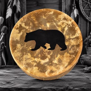 May include: A round drum featuring a mottled tan and brown surface with a solid black silhouette of a walking bear centered on the front. The drum head has a textured, organic appearance with varying shades of light and dark earth tones.