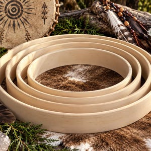 May include: A set of four nested circular wooden drum hoops with a smooth, light-colored finish. The hoops are arranged concentrically, decreasing in size from the outer ring to the center, showcasing a clean, unfinished wood grain texture.