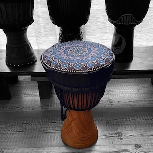 May include: A wooden djembe drum with a patterned blue and purple top. The drum's body is a light brown color, and the top has a dark blue background with a circular design of blue and white dots. Other black drums are in the background.