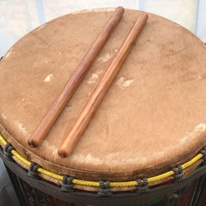 May include: A brown leather drum with a yellow rope border and two wooden drumsticks.