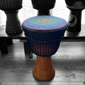 May include: A wooden djembe drum with a dark blue top featuring a colorful geometric design. The drum has a natural wood base and a dark blue body. The background shows other drums and a wooden bench.