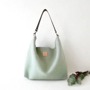 May include: A light green felt tote bag with a brown leather strap and a small leather patch on the front.