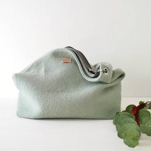 May include: A light green felt tote bag with a brown leather strap and a silver metal buckle. The bag is sitting on a white surface with a sprig of green leaves.