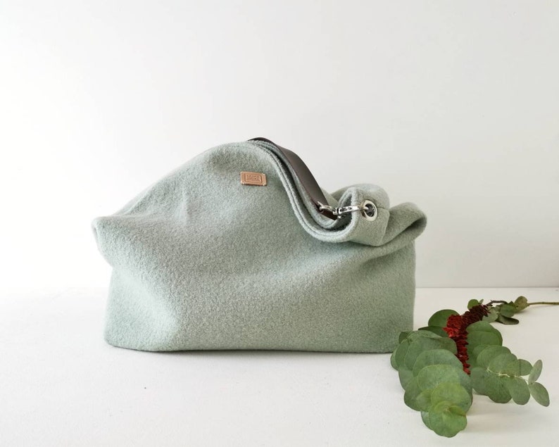 May include: A light green felt tote bag with a brown leather strap and a silver metal buckle. The bag is sitting on a white surface with a sprig of green leaves.