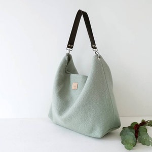 May include: A light green felt tote bag with a brown leather strap and silver hardware. The bag has a simple, minimalist design.