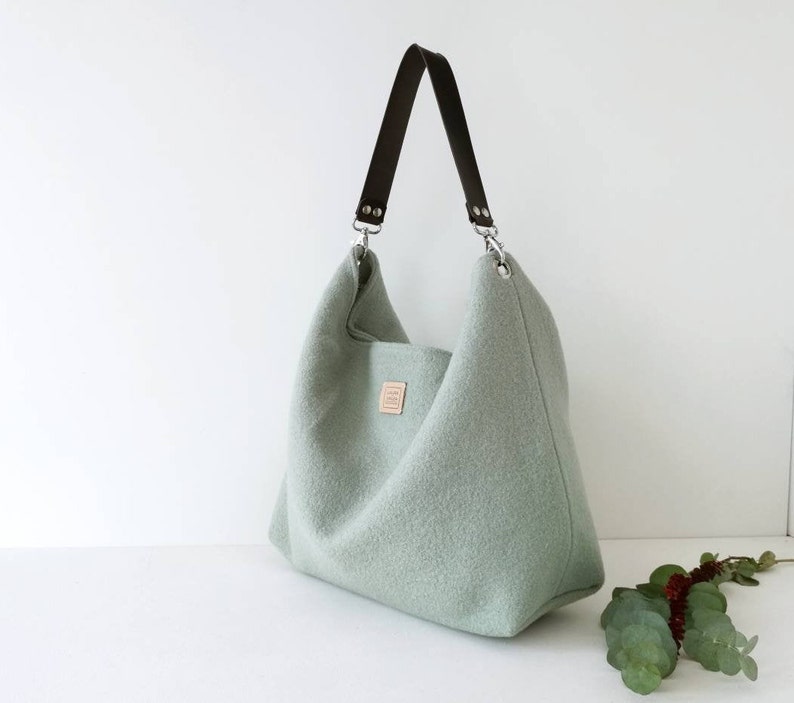 May include: A light green felt tote bag with a brown leather strap and silver hardware. The bag has a simple, minimalist design.