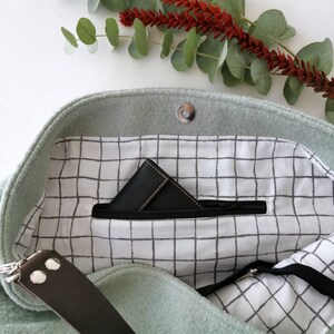 May include: A light green felt tote bag with a zippered pocket and a black and white grid pattern lining. The bag has a brown leather strap.