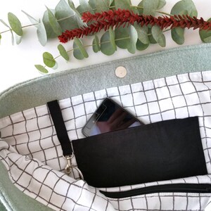 May include: A light green felt tote bag with a black zippered pocket and a black phone inside. The bag has a white and gray grid pattern lining.