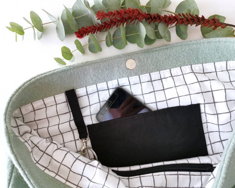 May include: A light green felt tote bag with a black zippered pocket and a black phone inside. The bag has a white and gray grid pattern lining.