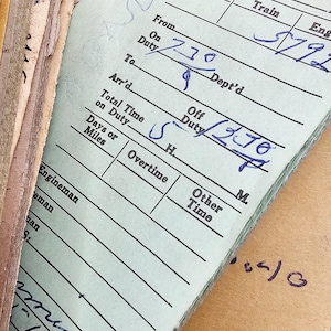 May include: Close-up of a stack of vintage train records. The top sheet is light green with handwritten entries in blue ink, including times and dates. A paper clip secures the top sheet. The records are aged and show signs of wear.