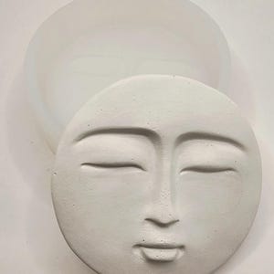 May include: A white, circular mold with a serene face design. The face has closed eyes, a small nose, and a slightly open mouth. The mold is made of a flexible material, with a matching, larger, circular base.