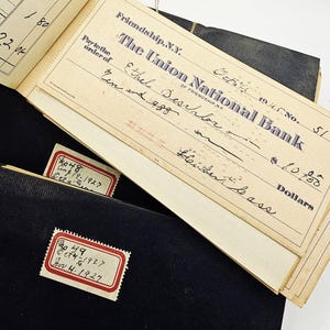 May include: An open vintage checkbook from The Union National Bank of Friendship, N.Y. dated October 1945, with a check made out to Ethel Deschlor for $10.00. Two black ledger books with attached labels are also visible.