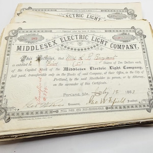 May include: A stack of antique Middlesex Electric Light Company stock certificates. The certificates are cream-colored with ornate black and red text and decorative borders. The text includes the company name, certificate number, and shareholder information.
