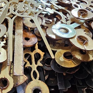 May include: A collection of vintage clock parts, including hands, gears, and decorative elements. The components are made of brass and other metals, with varying shades of gold and bronze. The parts are intricately designed and show signs of age.
