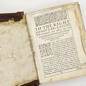 May include: An open antique book with a worn leather cover. The aged pages display ornate text and decorative elements, including a large initial letter and floral designs. The title reads "TO THE RIGHT HONOVRABLE, RIGHT VVorſhipfull..."