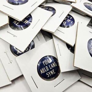 May include: A close-up of a pile of white card stock squares with blue and white button pins inside. The pins have various sayings on them, including "Let's Take a Joy Ride", "For Auld Lang Syne", and "I'm On My Way".