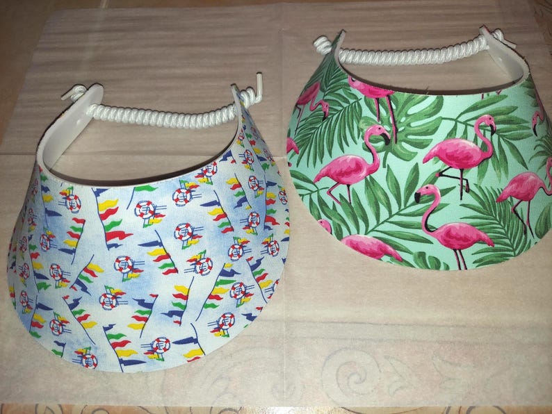 May include: Two sun visors with white rope headbands. The left visor features a light blue background with a pattern of colorful pennant flags and life preservers. The right visor has a light green background with a pattern of pink flamingos and green palm leaves.