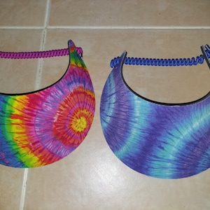 May include: Two sun visors with tie-dye patterns. The left visor features a vibrant rainbow spiral design with a purple cord strap. The right visor displays a blue and purple spiral pattern with a matching blue cord strap for secure placement.