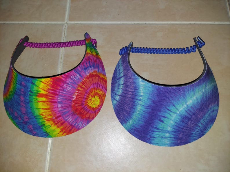 May include: Two sun visors with tie-dye patterns. The left visor features a vibrant rainbow spiral design with a purple cord strap. The right visor displays a blue and purple spiral pattern with a matching blue cord strap for secure placement.