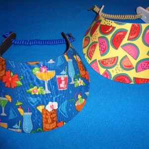 May include: Two sun visors with adjustable headbands. One visor features a blue background with a pattern of tropical cocktails, tiki statues, and flowers. The other visor has a yellow background with a repeating pattern of watermelon slices.