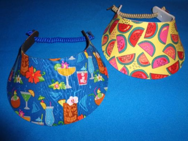 May include: Two sun visors with adjustable headbands. One visor features a blue background with a pattern of tropical cocktails, tiki statues, and flowers. The other visor has a yellow background with a repeating pattern of watermelon slices.