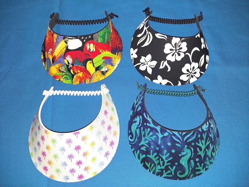May include: Four sun visors with coil headbands. Top left features a colorful parrot and toucan print. Top right has a black background with white hibiscus flowers. Bottom left is white with small rainbow palm trees. Bottom right shows a blue sea life pattern.