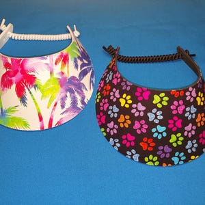 May include: Two sun visors with coiled elastic headbands. The left visor features a white background with a colorful palm tree print in shades of pink, purple, and green. The right visor has a black background with a repeating pattern of multicolored paw prints.