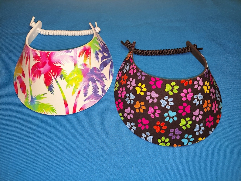 May include: Two sun visors with coiled elastic headbands. The left visor features a white background with a colorful palm tree print in shades of pink, purple, and green. The right visor has a black background with a repeating pattern of multicolored paw prints.