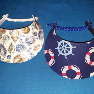 May include: Two sun visors. The left visor is white with a pattern of various brown and blue seashells. The right visor is dark blue with a pattern of light blue ship steering wheels and red and white life preservers. Both have coiled bands.