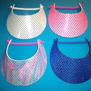 May include: Four sun visors with coiled elastic headbands. One is white with black polka dots, one is pink with black polka dots, one is blue with a glittery texture, and one features a multi-colored swirling pattern with a glittery finish.