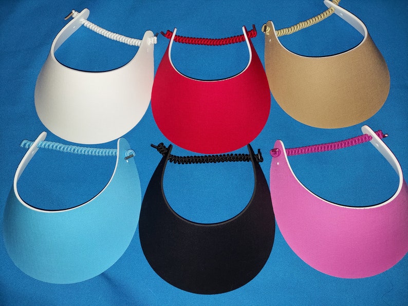 May include: A collection of six sun visors in white, red, tan, light blue, black, and pink. Each visor features a curved brim and a coiled, elasticized strap that connects the two sides, providing a flexible fit for various head sizes.