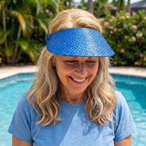 May include: A bright blue sun visor with a textured, glittery finish. The visor features a wide, curved brim designed to provide shade, with a small blue loop detail on the side. The material has a consistent, sparkling appearance across the surface.