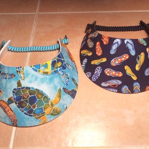 May include: Two sun visors with adjustable coil bands. The left visor features a light blue background with a colorful sea turtle pattern. The right visor has a dark navy blue background with a repeating pattern of multi-colored flip-flop sandals.