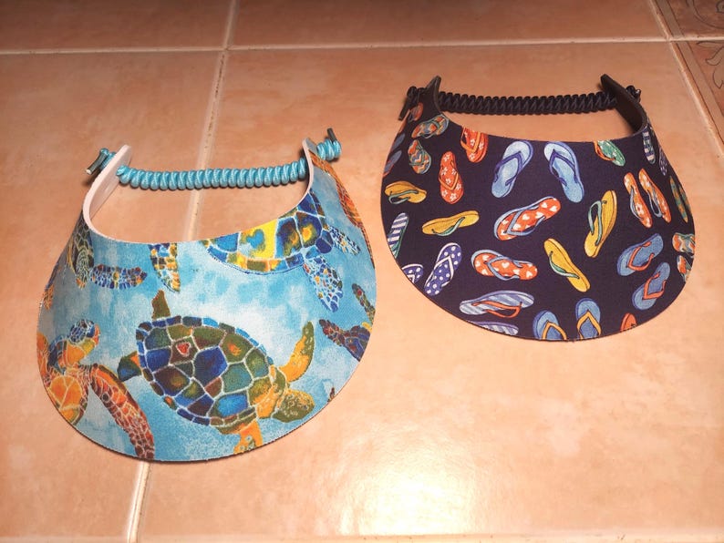 May include: Two sun visors with adjustable coil bands. The left visor features a light blue background with a colorful sea turtle pattern. The right visor has a dark navy blue background with a repeating pattern of multi-colored flip-flop sandals.