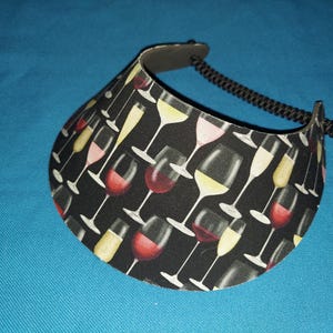 May include: A sun visor featuring a black background with a repeating pattern of various wine glasses filled with red, white, and pink wine. The visor has a curved brim and an adjustable black coiled cord strap attached to the sides for securing.