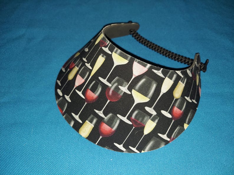 May include: A sun visor featuring a black background with a repeating pattern of various wine glasses filled with red, white, and pink wine. The visor has a curved brim and an adjustable black coiled cord strap attached to the sides for securing.