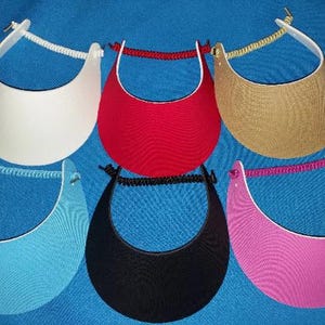 May include: Six sun visors in white, red, tan, light blue, black, and pink. Each visor features a curved brim and a white plastic headband connected by a coiled, color-coordinated elastic cord at the back for an adjustable fit.